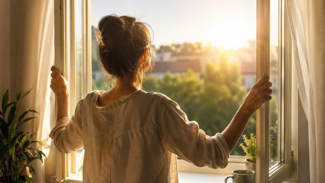 Vrouw opent ’s ochtends een raam en laat frisse lucht binnen als symbool voor nieuw perspectief en intuïtieve coaching